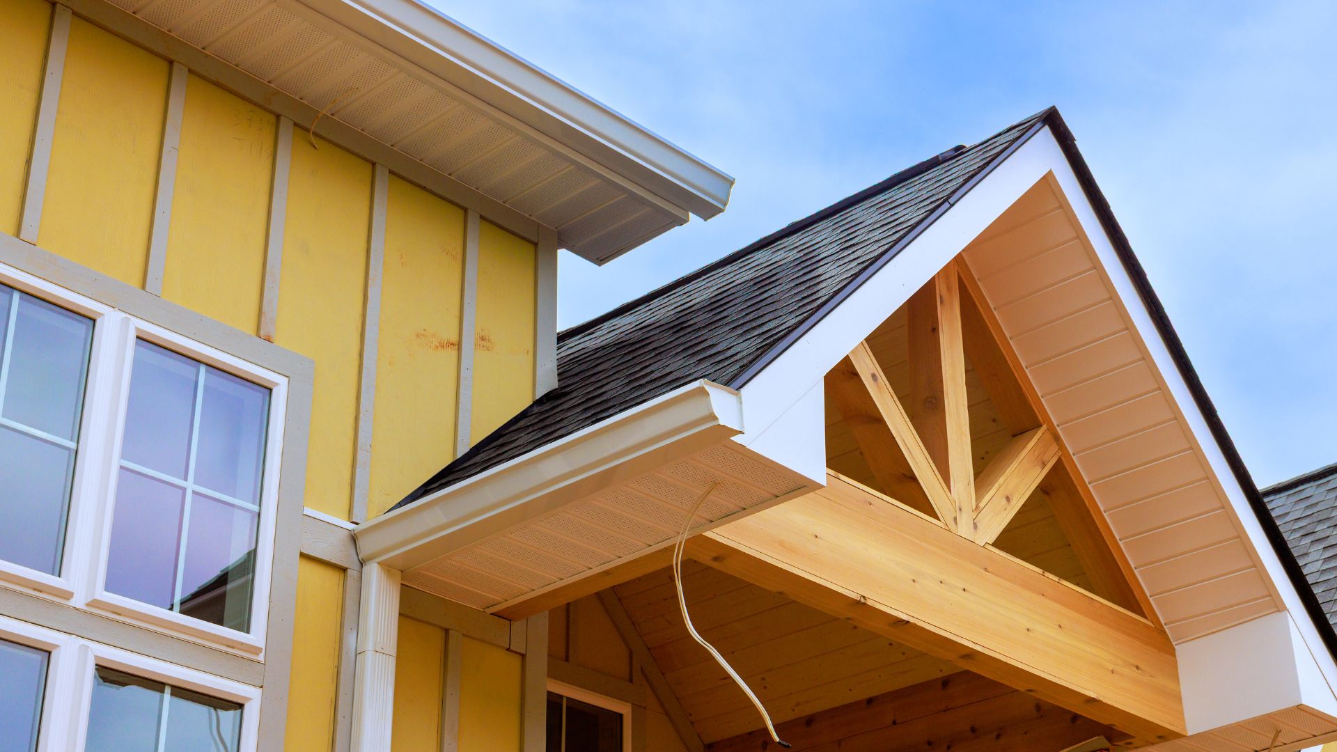 Close-up view of the upper section of a yellow house with white trim, showing large windows, a gabled roof, exposed wooden beams, and blue sky in the background.