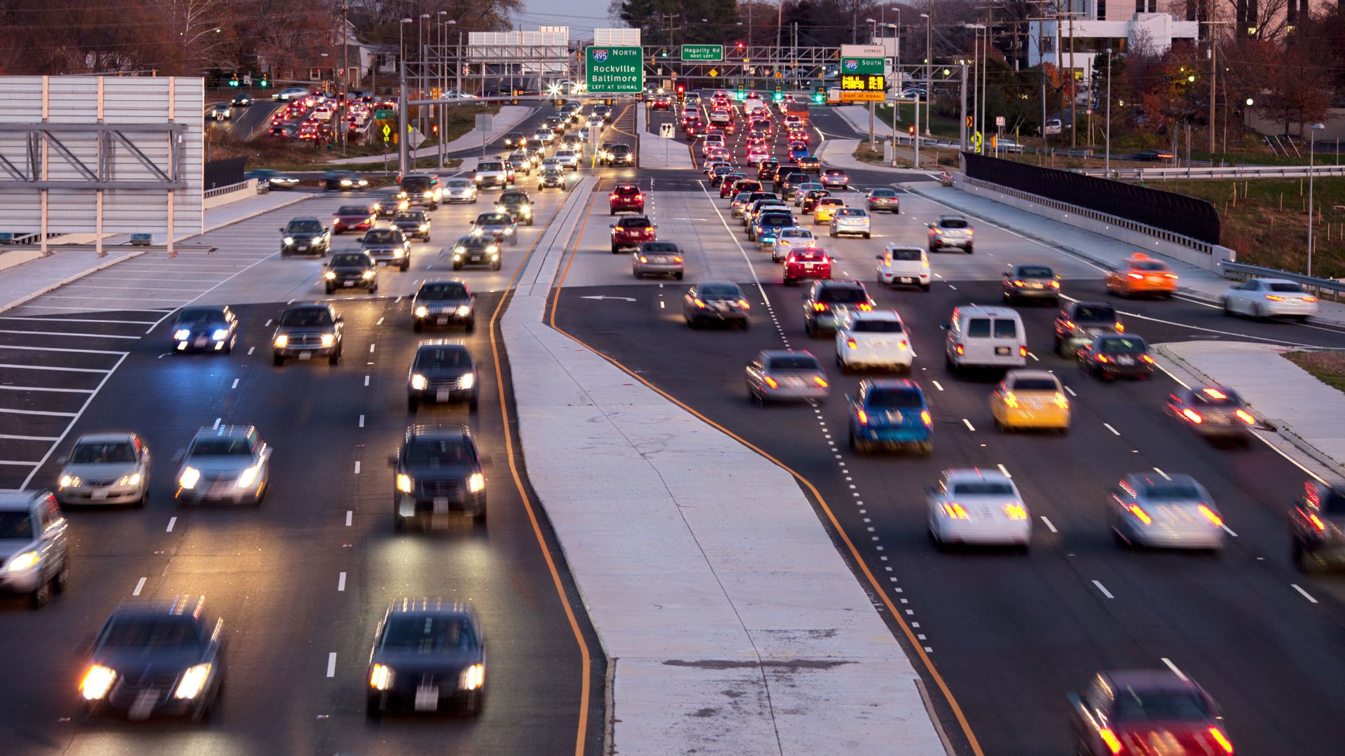 A busy multi-lane highway with heavy traffic flowing in both directions during dusk. Headlights and taillights of cars create streaks of light, and green highway signs are visible overhead. Trees and buildings line the road.