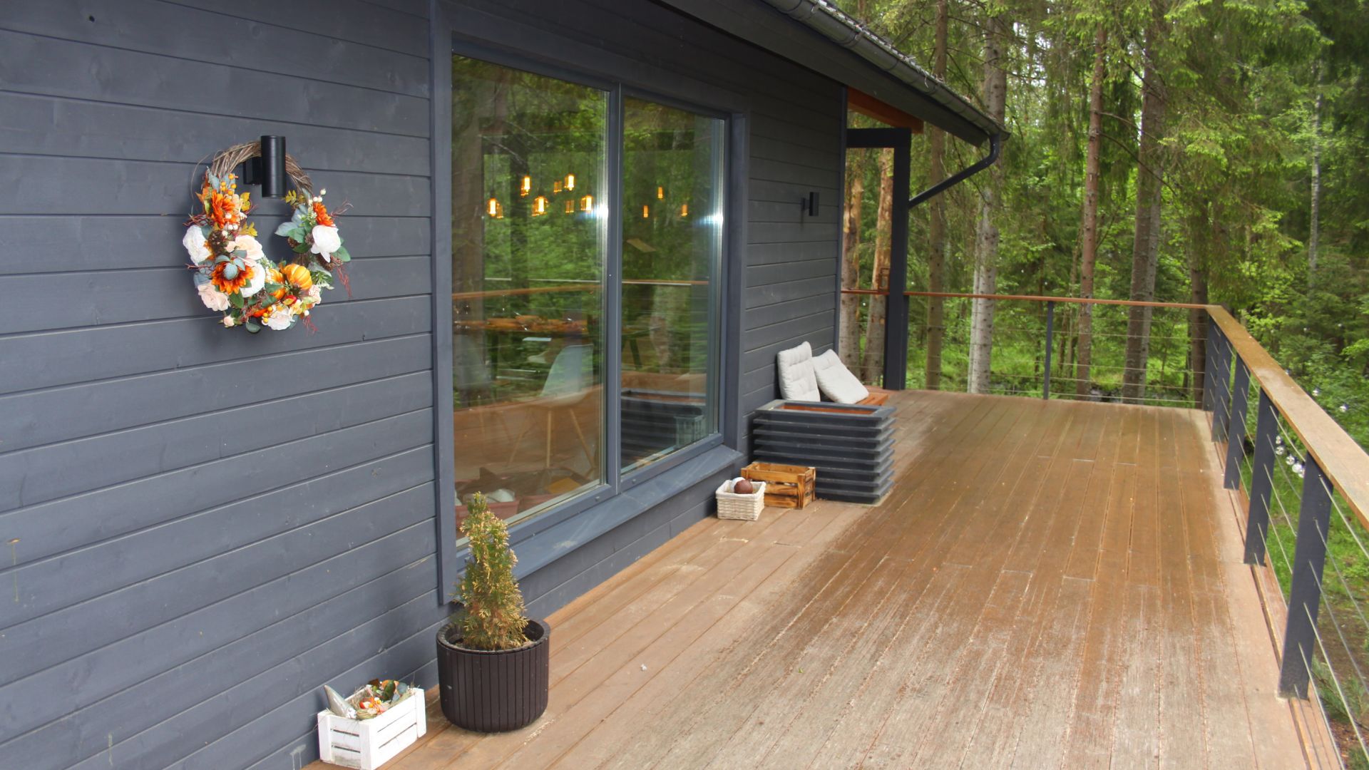 A wooden deck attached to a dark blue house with a large window, decorated with a floral wreath, potted plant, and stacked cushions. The deck overlooks a lush green forest.