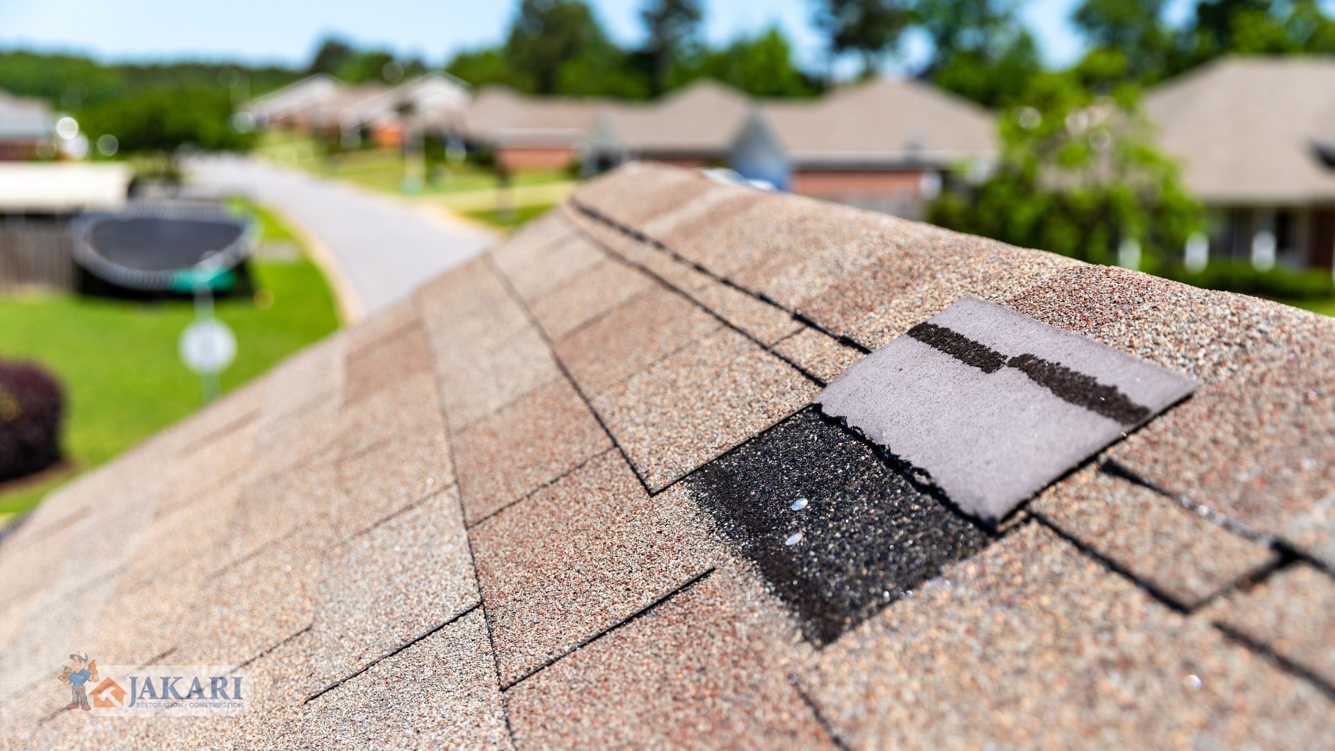 Close-up of a residential roof with a damaged or missing shingle, exposing the black underlayer, with a suburban neighborhood and green trees blurred in the background.